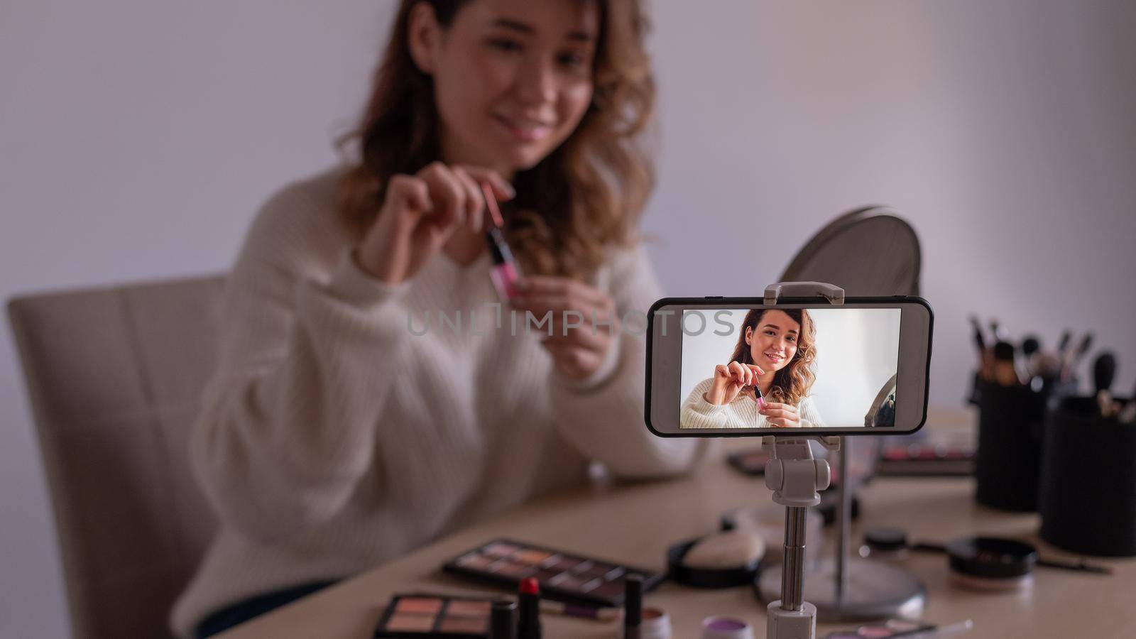 Young caucasian woman is recording an online make-up training video on her mobile. Video blogger girl applies makeup