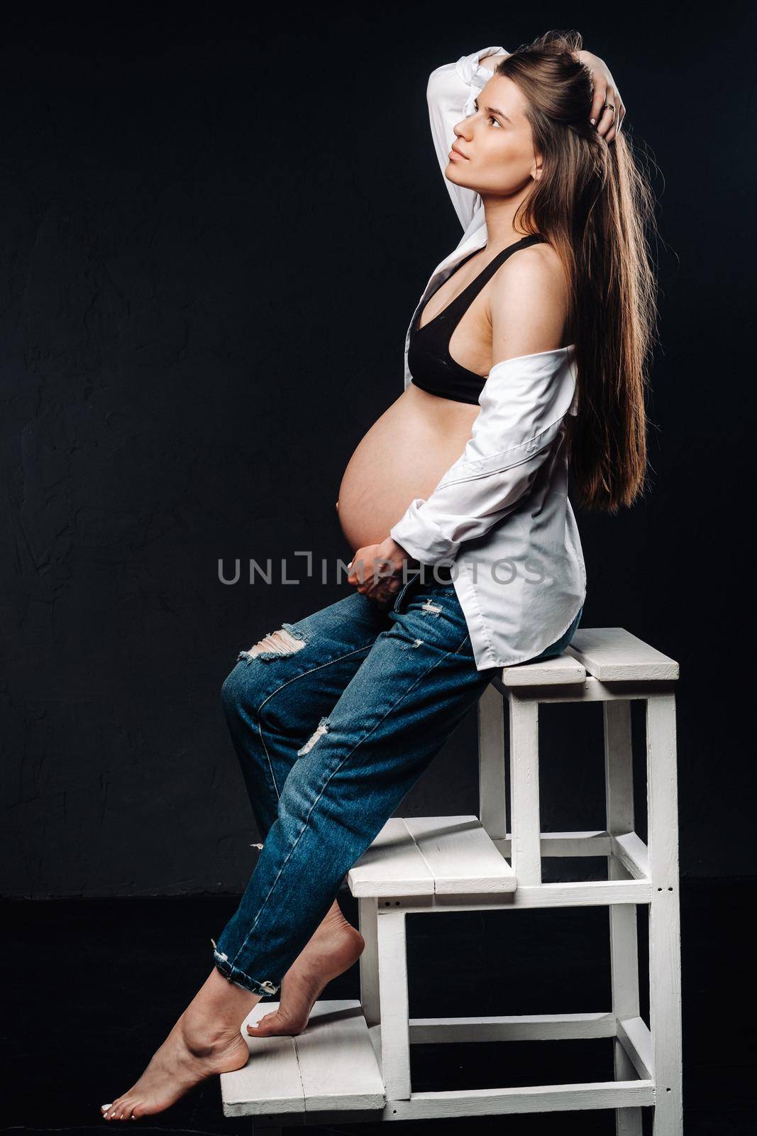 pregnant woman sitting on a ladder chair in a studio on a black background.