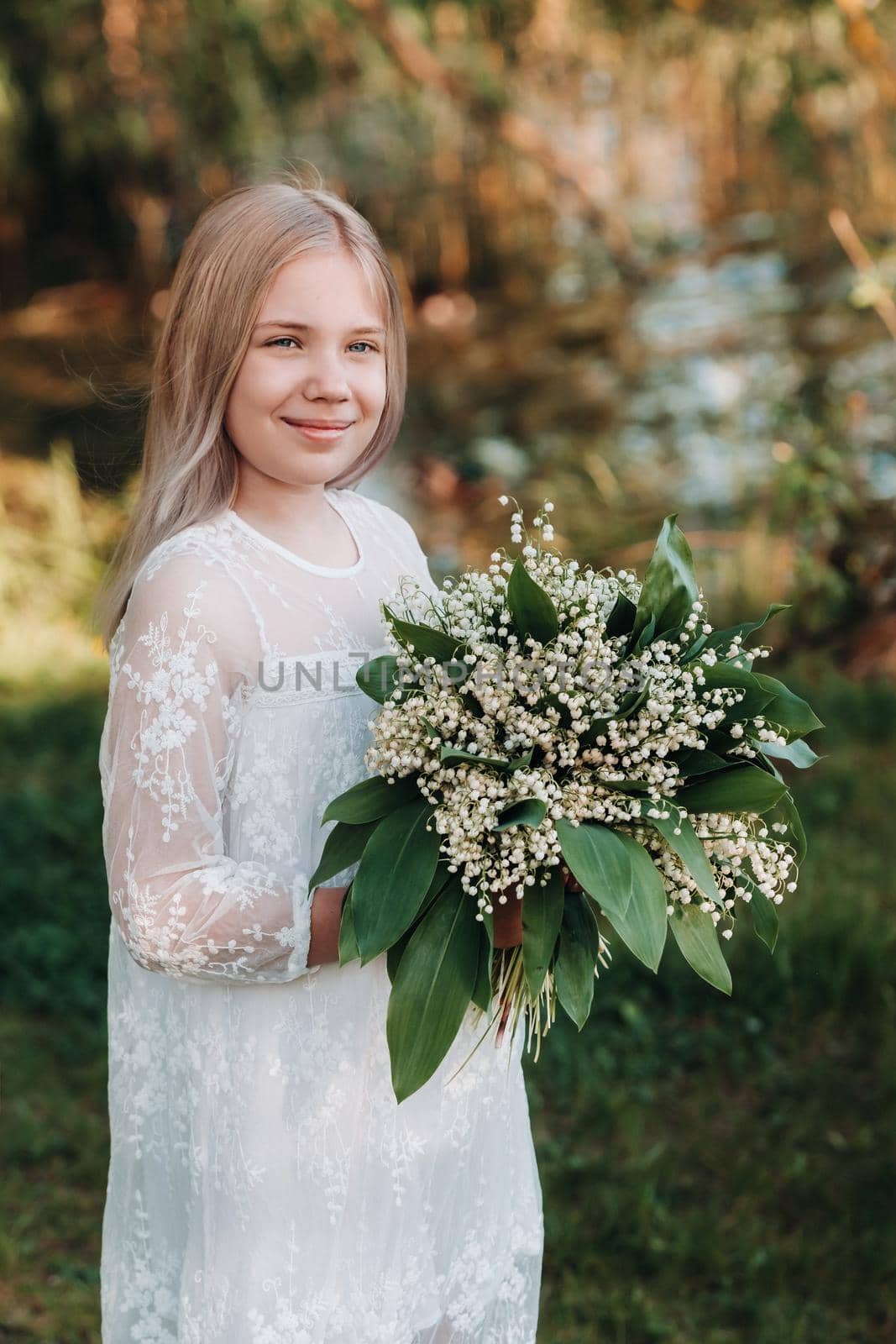 A beautiful nine-year-old blonde girl with long hair in a long white dress, holding a bouquet of lilies of the valley flowers, walking in nature in the Park.Summer, sunset