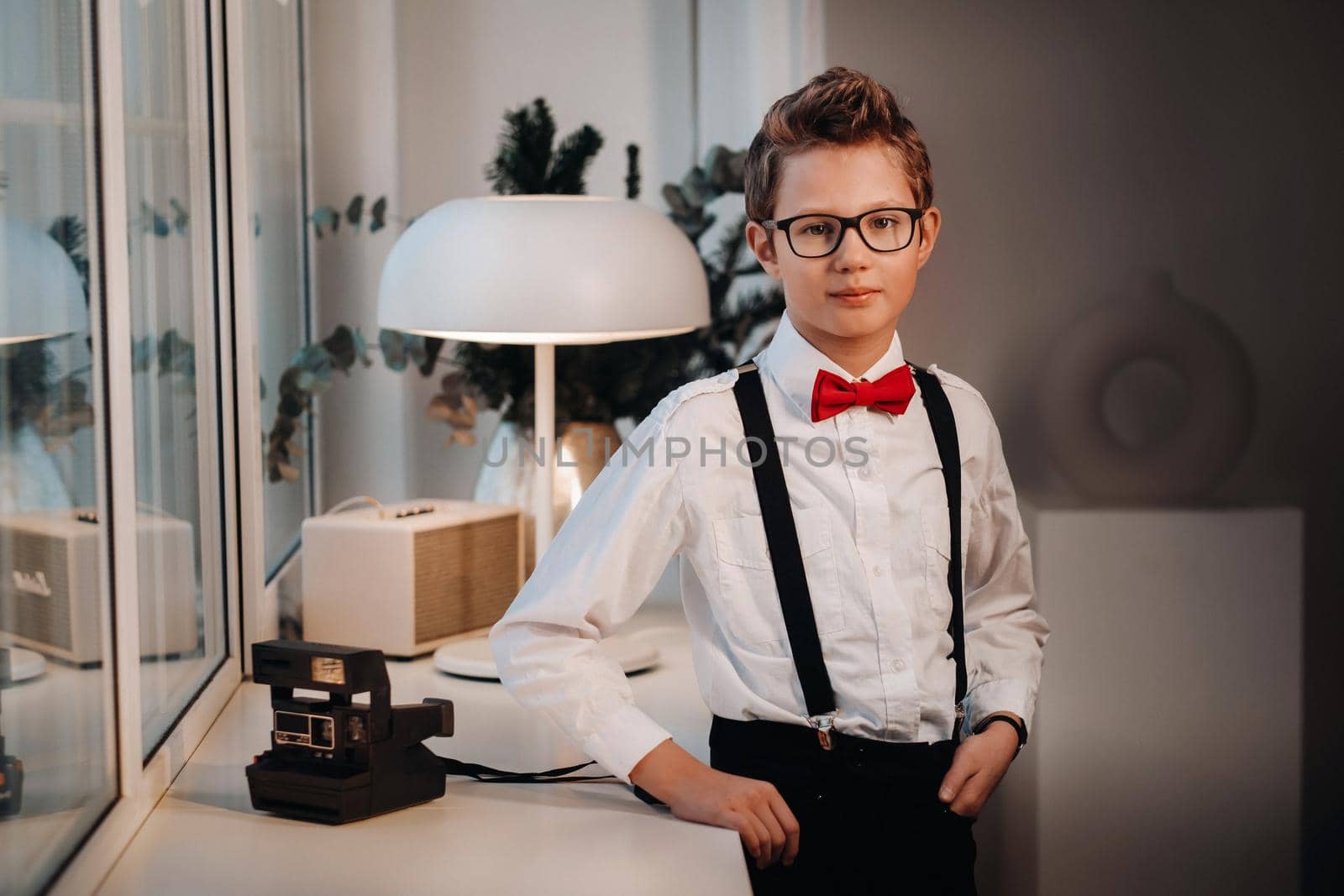 A boy in a shirt with a red bow tie and glasses stands at the window