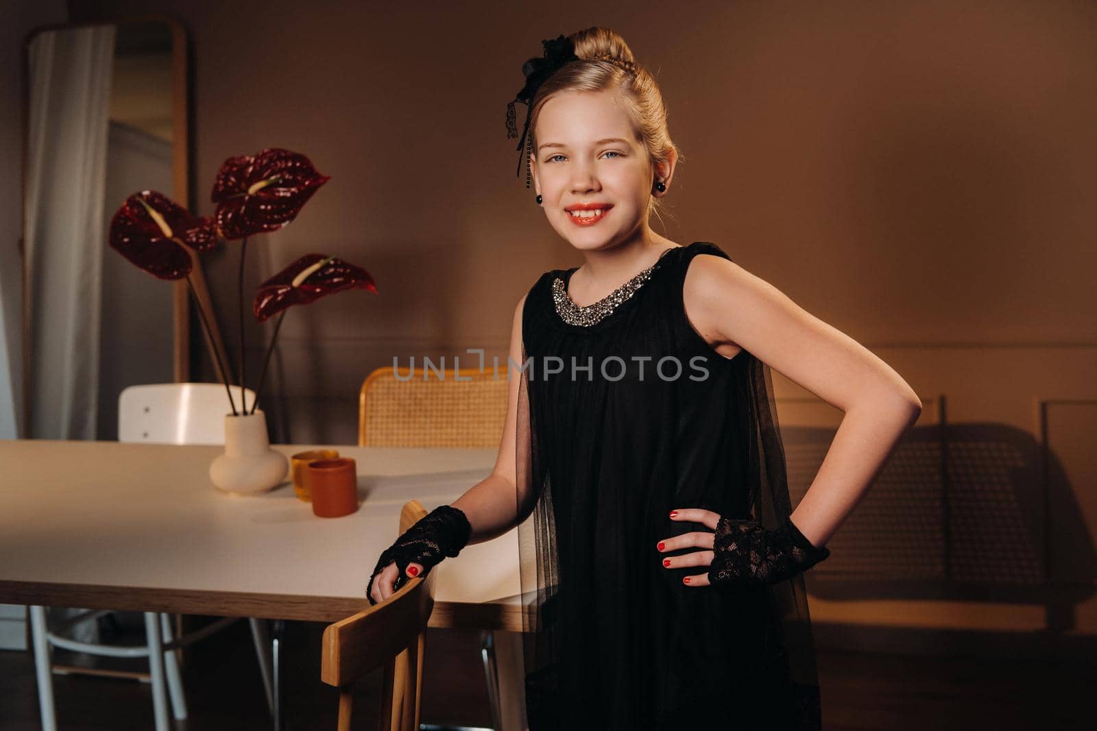 A stylish little girl in a black dress stands in the interior near the table.