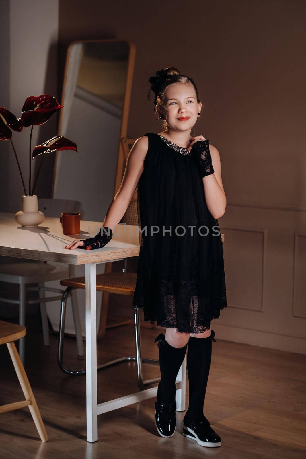 A stylish little girl in a black dress stands in the interior near the table.
