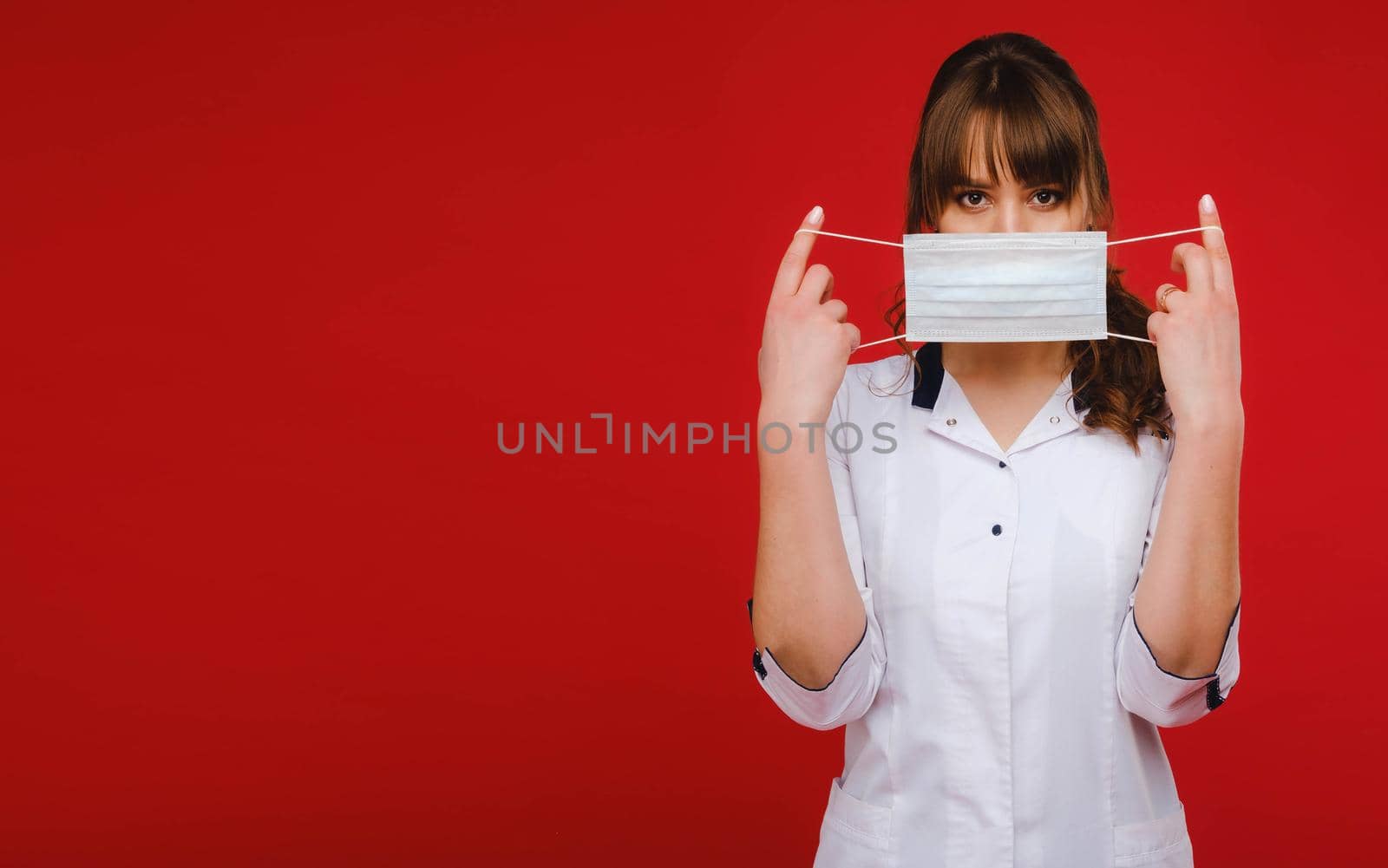A female doctor stands on a red background and holds a medical mask in her hands.