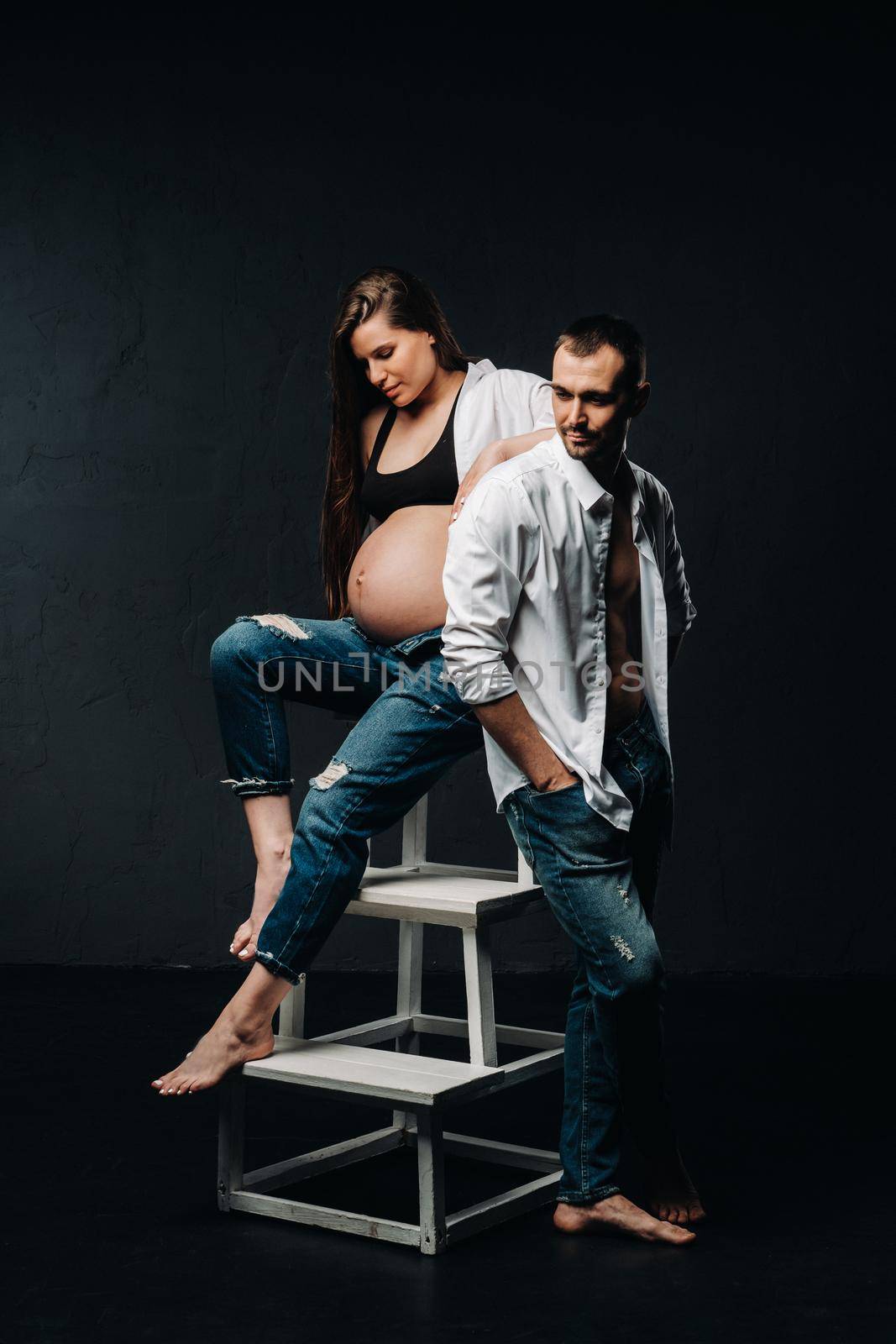 a pregnant woman and a man in a white shirt and jeans in a studio on a black background by Lobachad