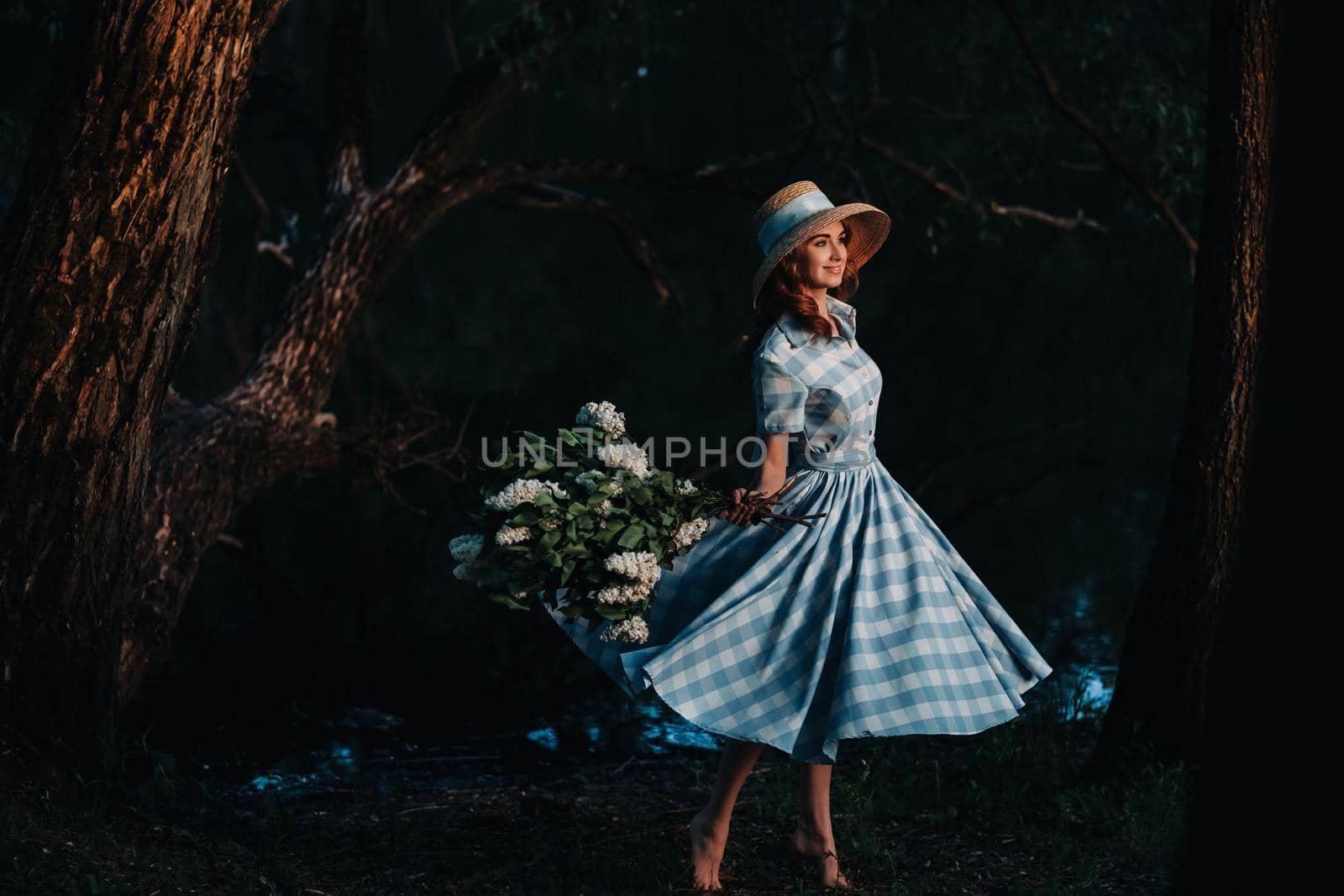 Portrait of a beautiful girl with long hair, a straw hat and a long summer dress with lilac flowers in the garden.