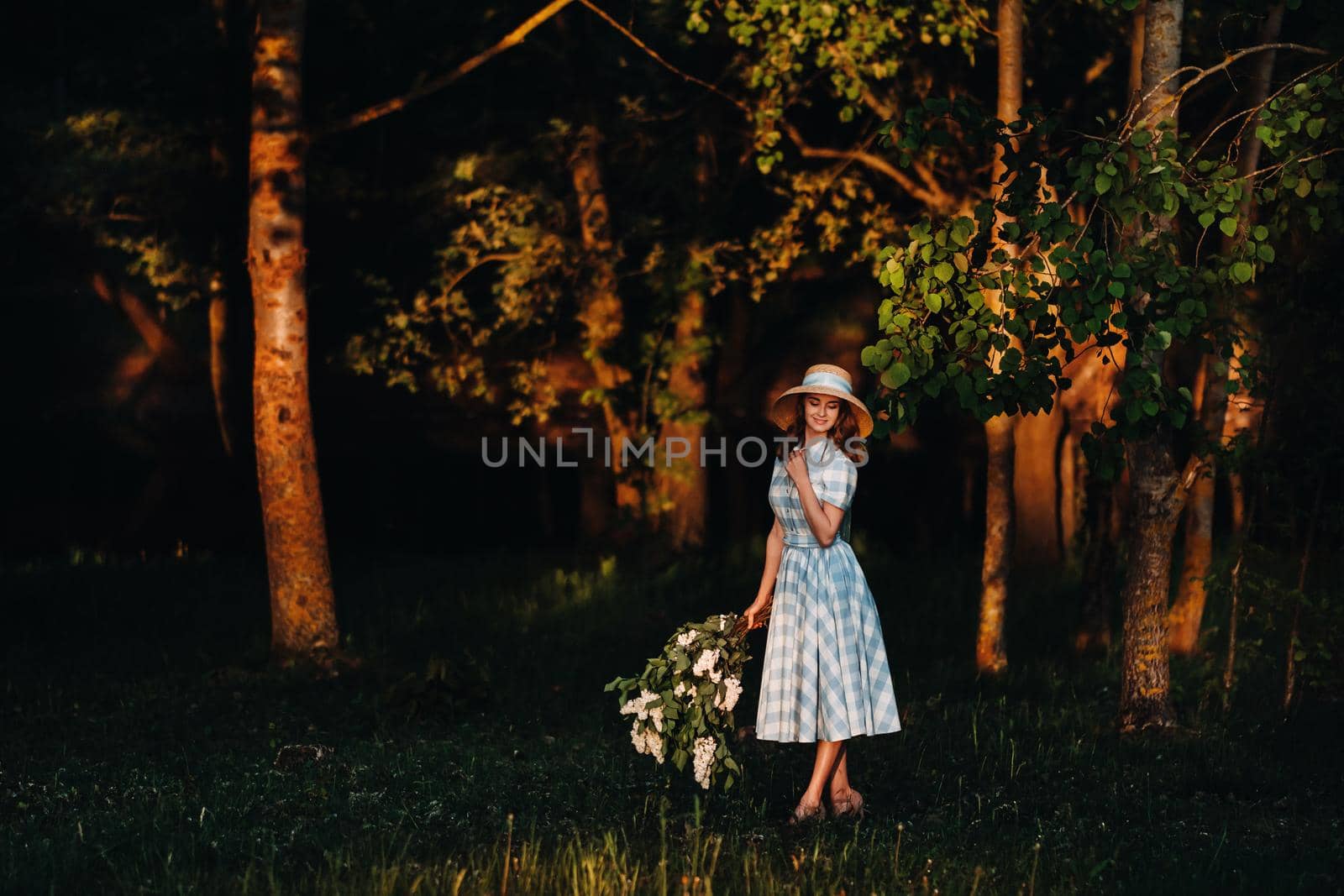 Portrait of a beautiful girl with long hair, a straw hat and a long summer dress with lilac flowers in the garden.