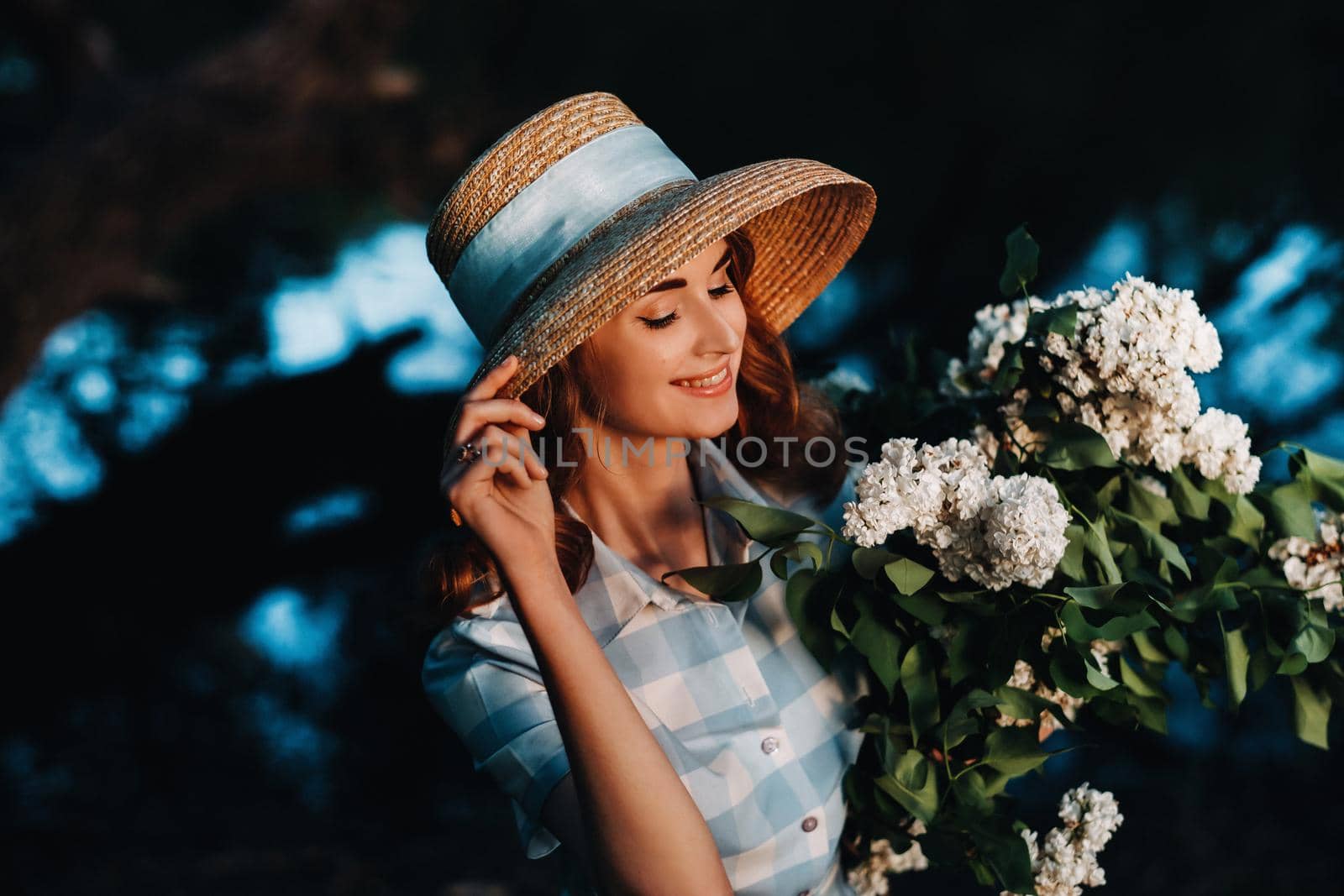 Portrait of a beautiful girl with long hair, a straw hat and a long summer dress with lilac flowers in the garden.