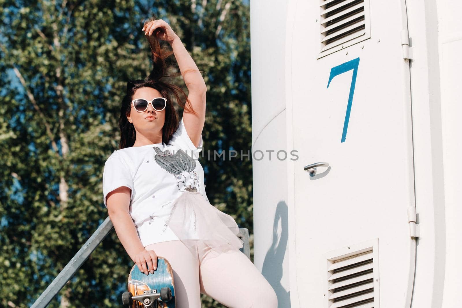 A girl in white clothes and glasses with a skate in her hands is photographed near large wind turbines in a field with trees.Modern woman with a riding Board in a field with windmills.