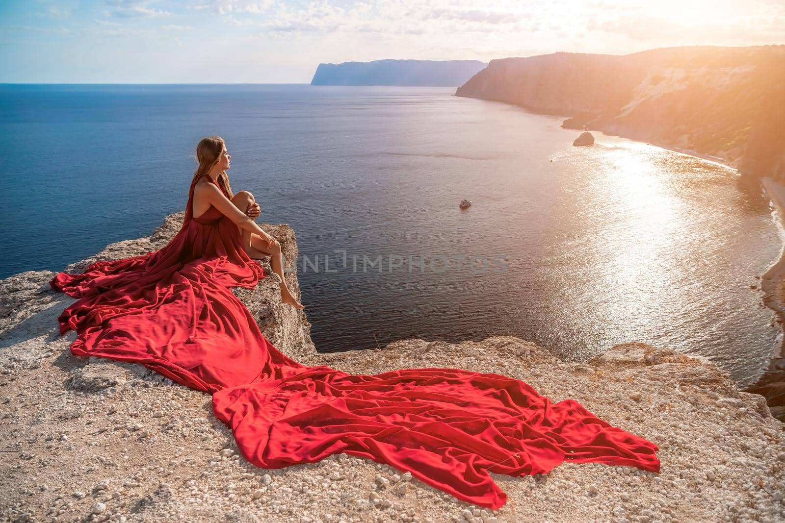 Side view a Young beautiful sensual woman in a red long dress posing on a rock high above the sea during sunrise. Girl on the nature on blue sky background. Fashion photo by panophotograph