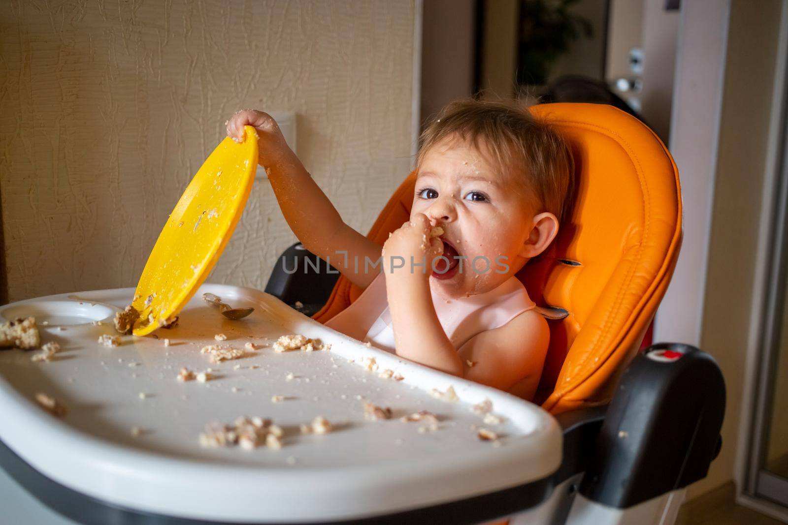 adorable baby plays with a plastic plate at the table. little child indulges in a baby chair after eating by Mariaprovector