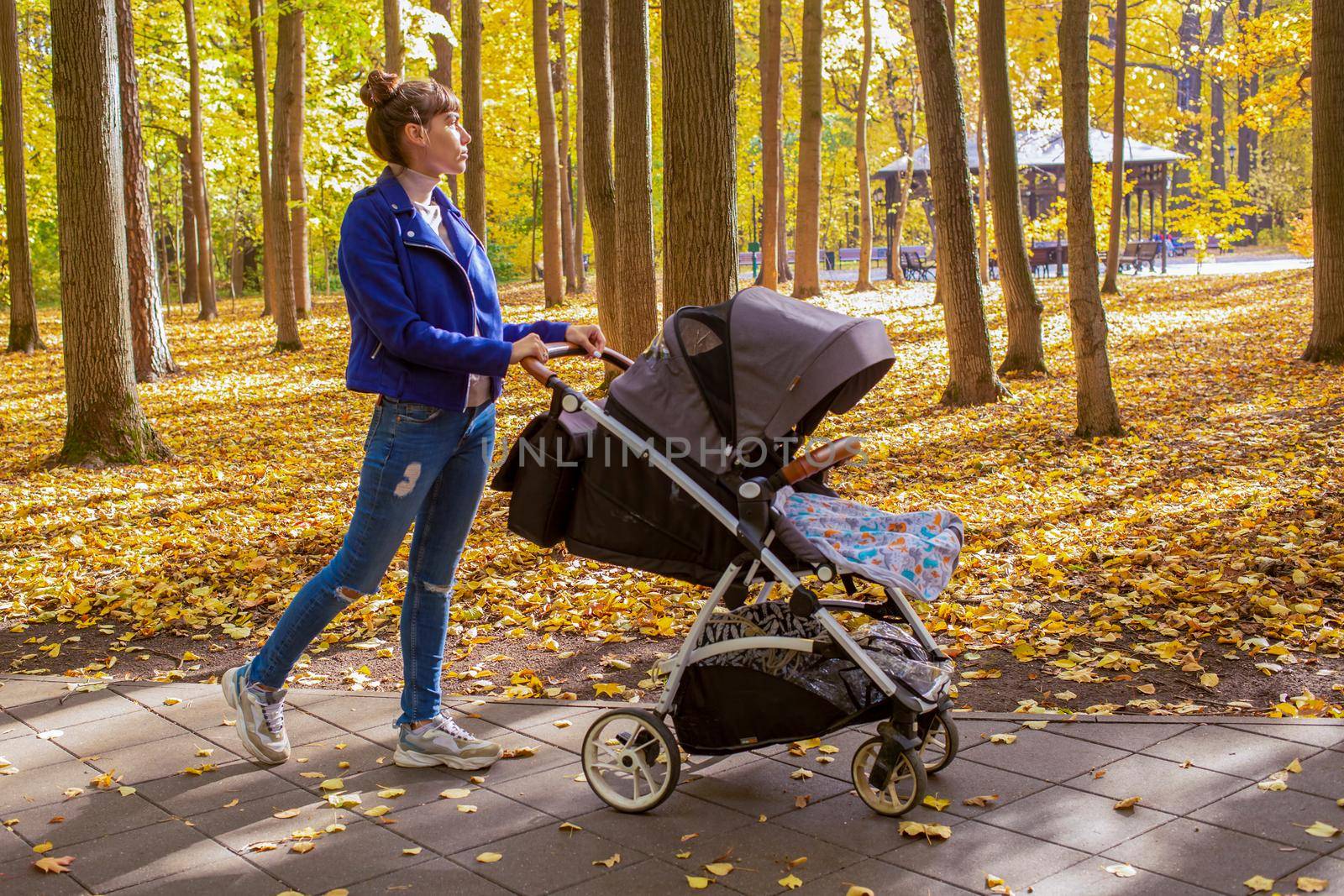 young mother goes with a stroller in the autumn park. a one-year-old infant is sleeping in a carriage. tired mom