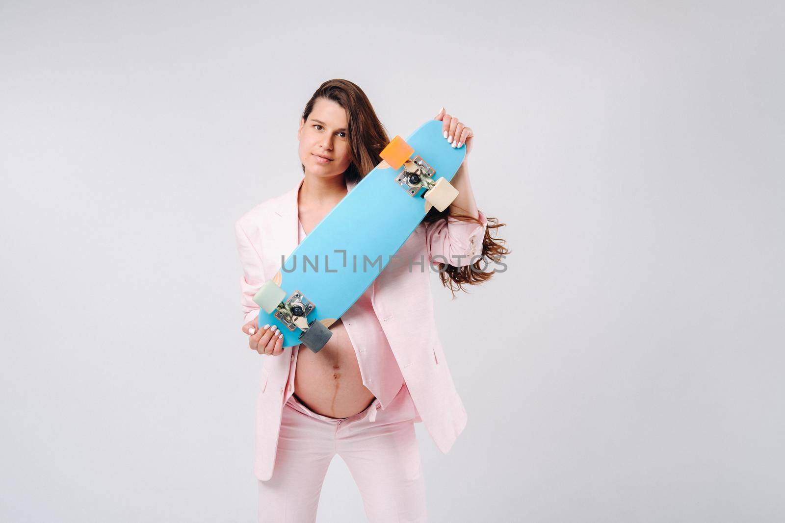 A pregnant girl in a pink suit with a skateboard in her hands stands on a gray background.