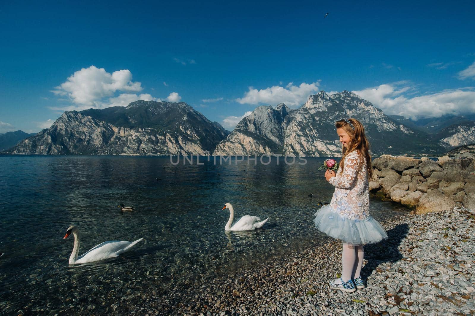 A little girl in a smart white dress walks along the embankment of lake Garda.A girl is photographed against the background of a mountain and lake in Italy.Torbole.