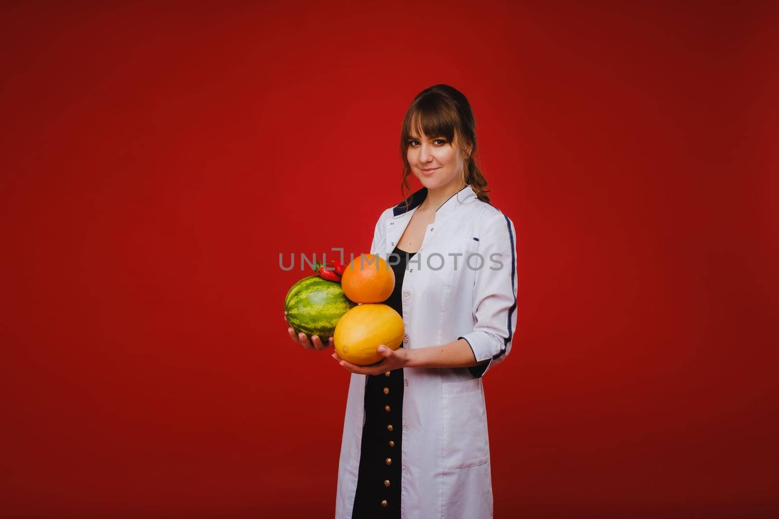 a female doctor nurse in a white coat with fruit in her hands poses on a red background, melon, watermelon, strawberry and grapefruit.