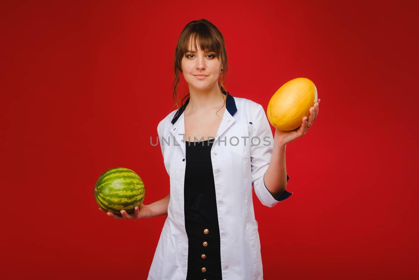 a female doctor nurse in a white coat with fruit in her hands poses on a red background, melon, watermelon