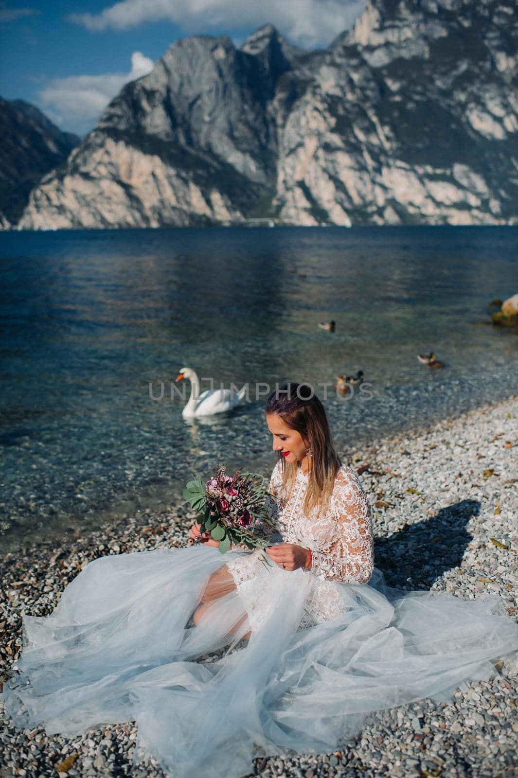 a girl in a smart white dress is sitting on the embankment of lake Garda.A woman is photographed against the background of a mountain and lake in Italy.Torbole.