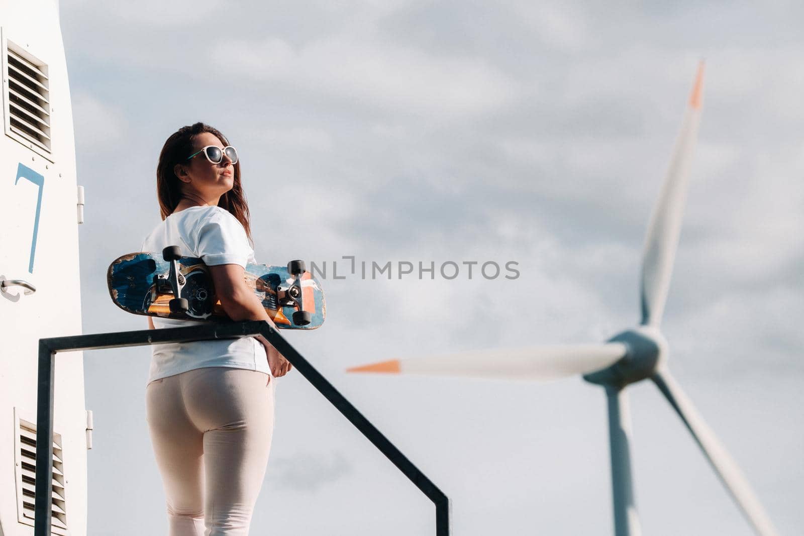 A girl in white clothes and glasses with a skate in her hands is photographed near large wind turbines in a field with trees.Modern woman with a riding Board in a field with windmills.