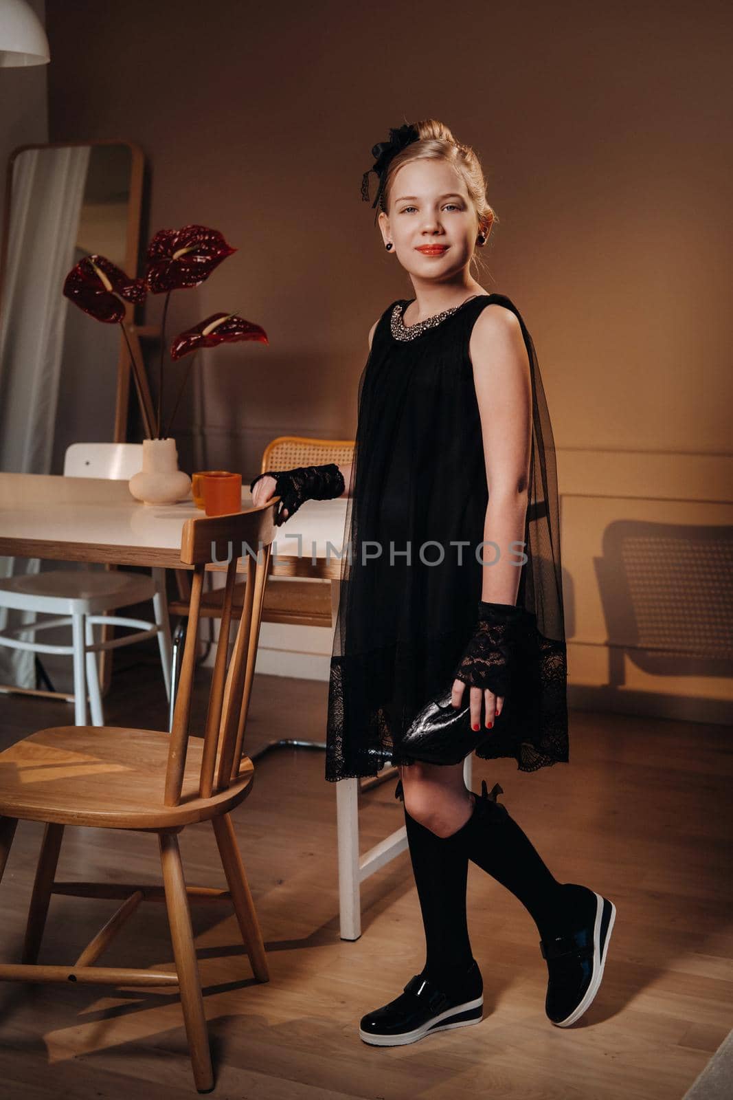 A stylish little girl in a black dress stands in the interior near the table by Lobachad