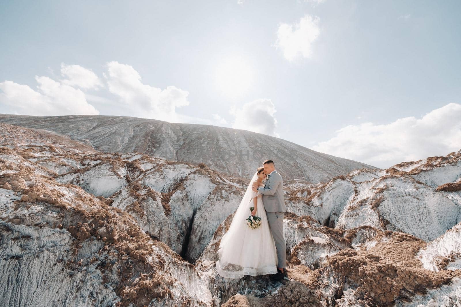 A beautiful couple of lovers posing in a white salt mountain. A young woman in a stylish wedding dress and a beautiful stylish man in a gray suit. The concept of the wedding day. by Lobachad