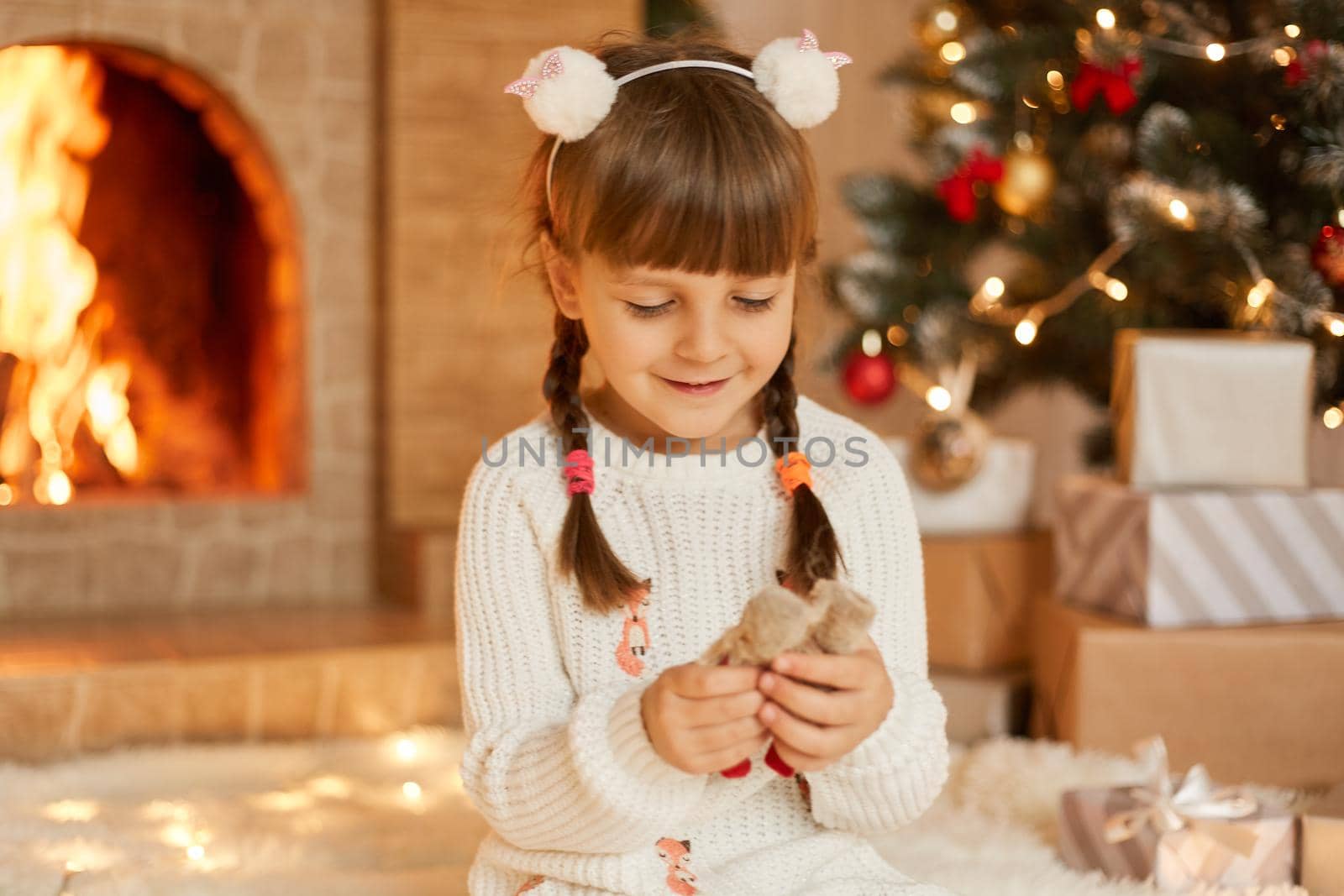 Charming kid girl playing with toy indoors, sitting on floor in room with Christmas decorations, celebration New Year, female child looking at her toy with smile, wearing white sweater.