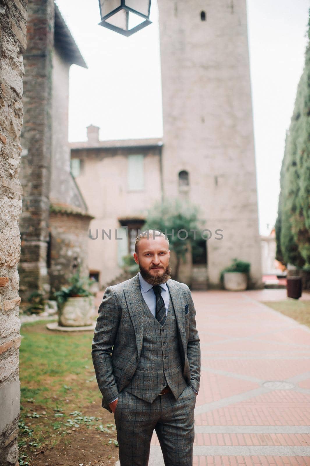 A man with a beard in a strict grey three-piece suit with a tie in the old town of Sirmione, a Stylish man in a grey suit in Italy by Lobachad