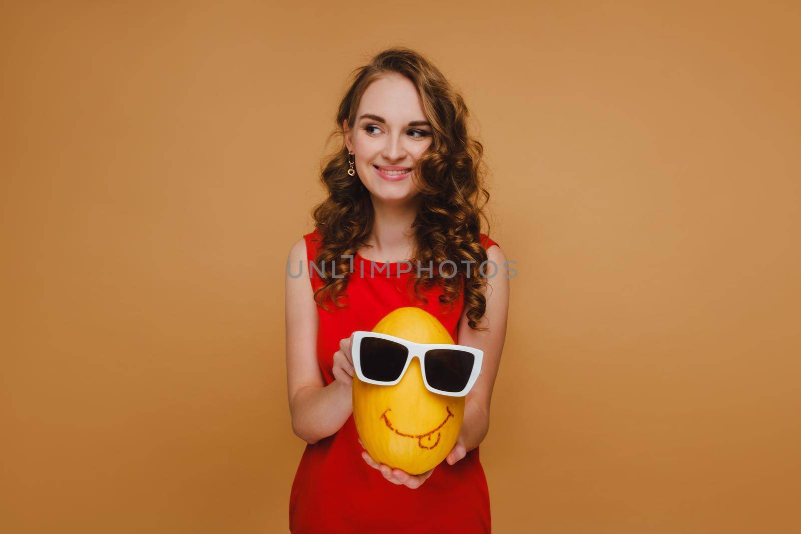 beautiful young girl holds hands melon. The melon is wearing glasses and an emoticon is drawn. Girl in a red dress.
