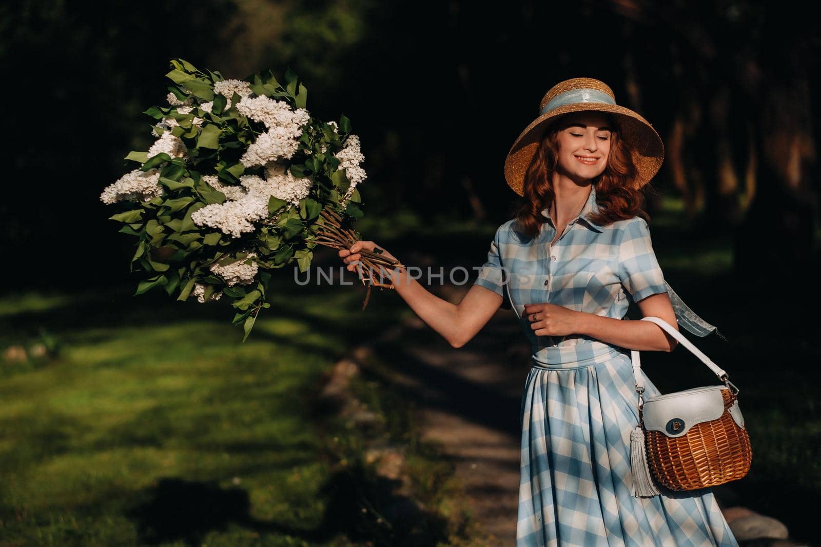 Portrait of a beautiful girl with long hair, a straw hat and a long summer dress with lilac flowers in the garden.