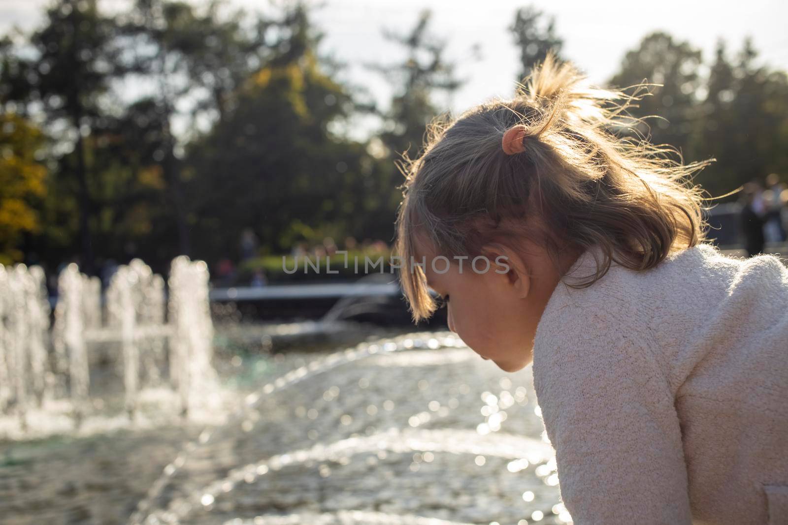adorable toddler girl looks at the pond with fountains in the park on a sunny day. weekend family walk. spending time with children by Mariaprovector