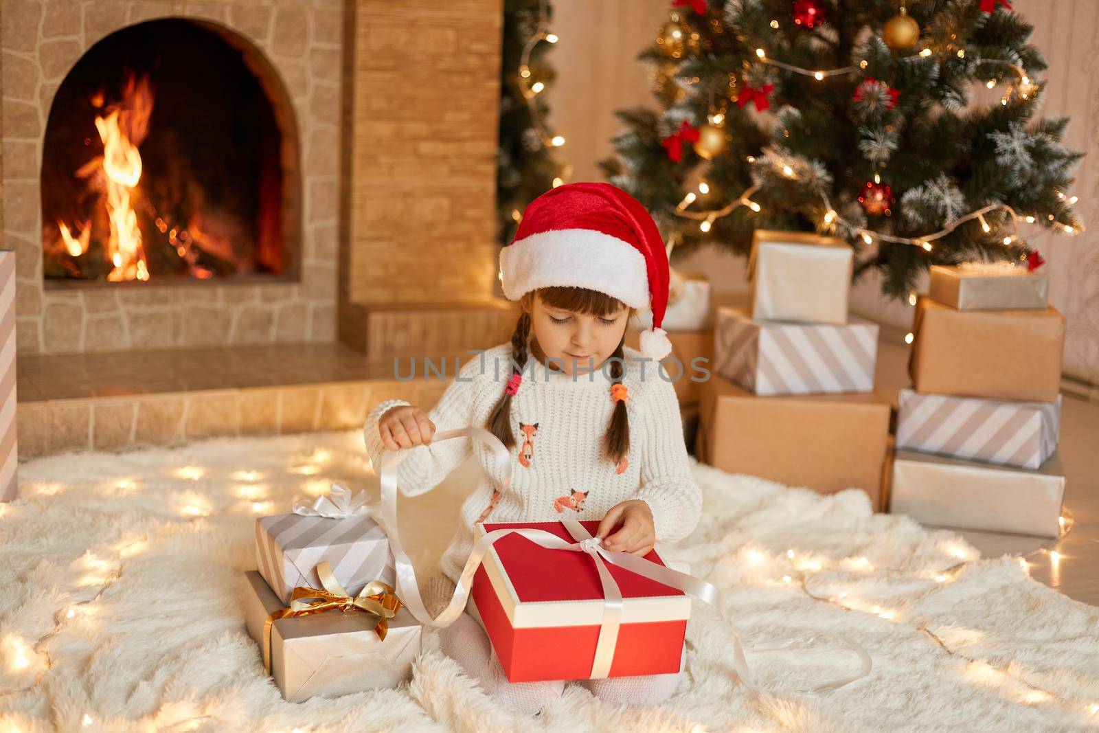 Cute female kid with pigtails sitting on floor and pulling ribbon from her present box, opening her gift, wearing pullover and santa hat, posing with fireplace and Christmas tree on background.