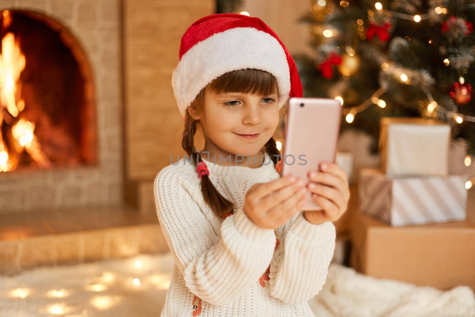 New Year's portrait of little cute girl against background of Christmas tree and fireplace, child holding mobile phone, has video call and congratulating, somebody, wearing festive red hat.