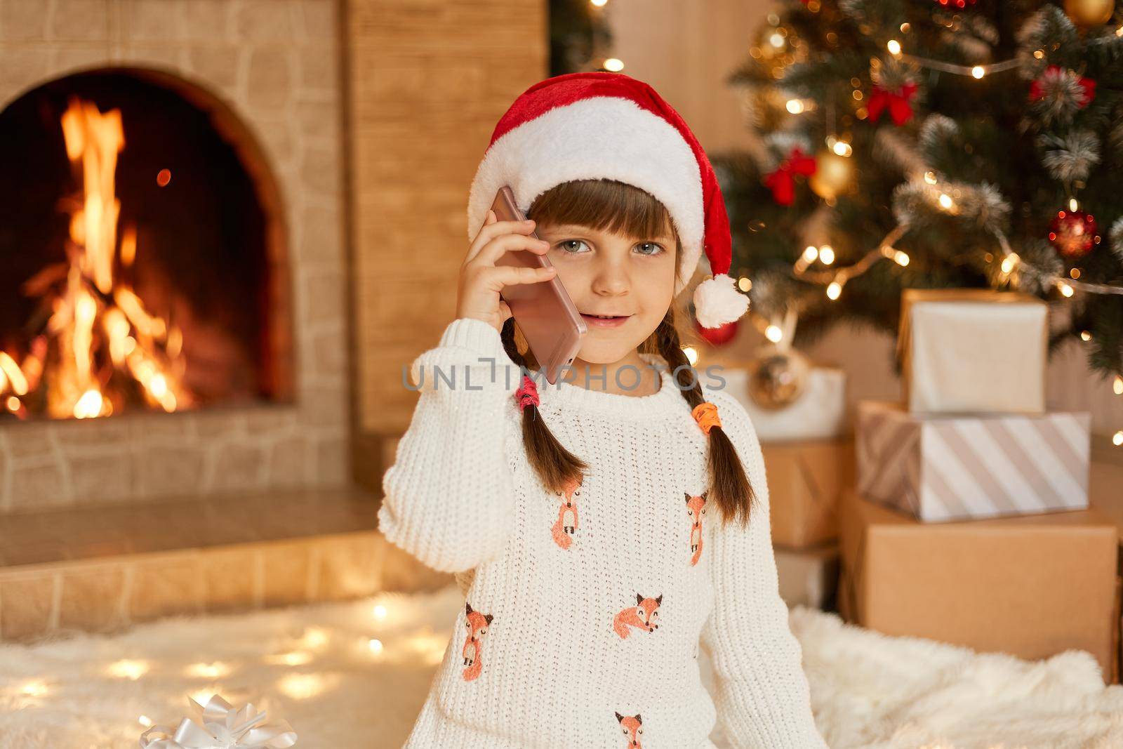 Attractive child wearing white pullover and santa claus hat talking phone in festive room, posing with fir tree, gift boxes and fireplace on background, looks at camera.