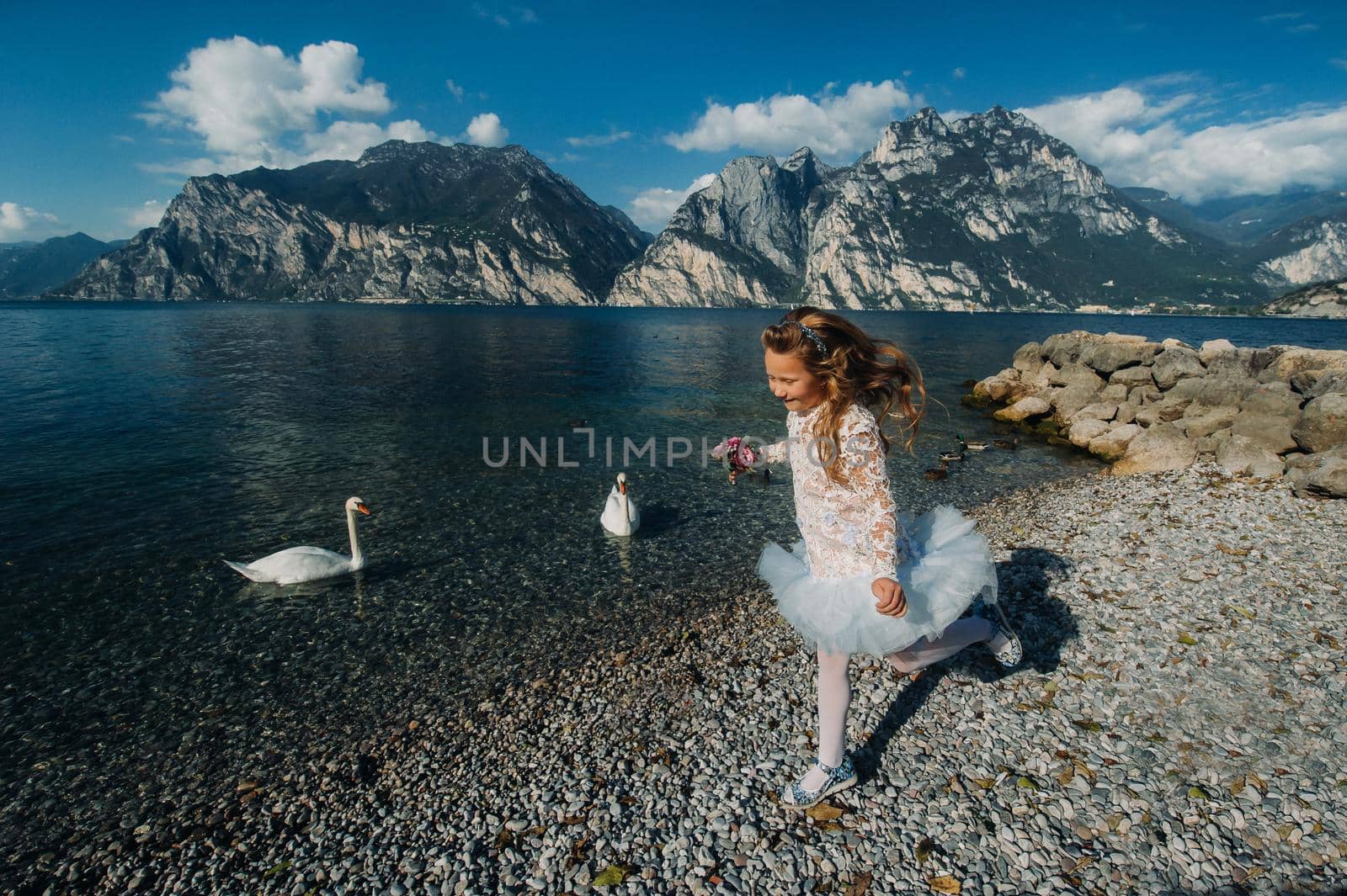 A little girl in a smart white dress walks along the embankment of lake Garda.A girl is photographed against the background of a mountain and lake in Italy.Torbole.