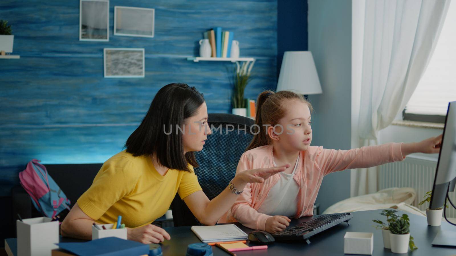 Parent and little girl doing homework together at desk and using computer with keyboard for online lesson. Mother helping daughter with school work and class tasks for remote education