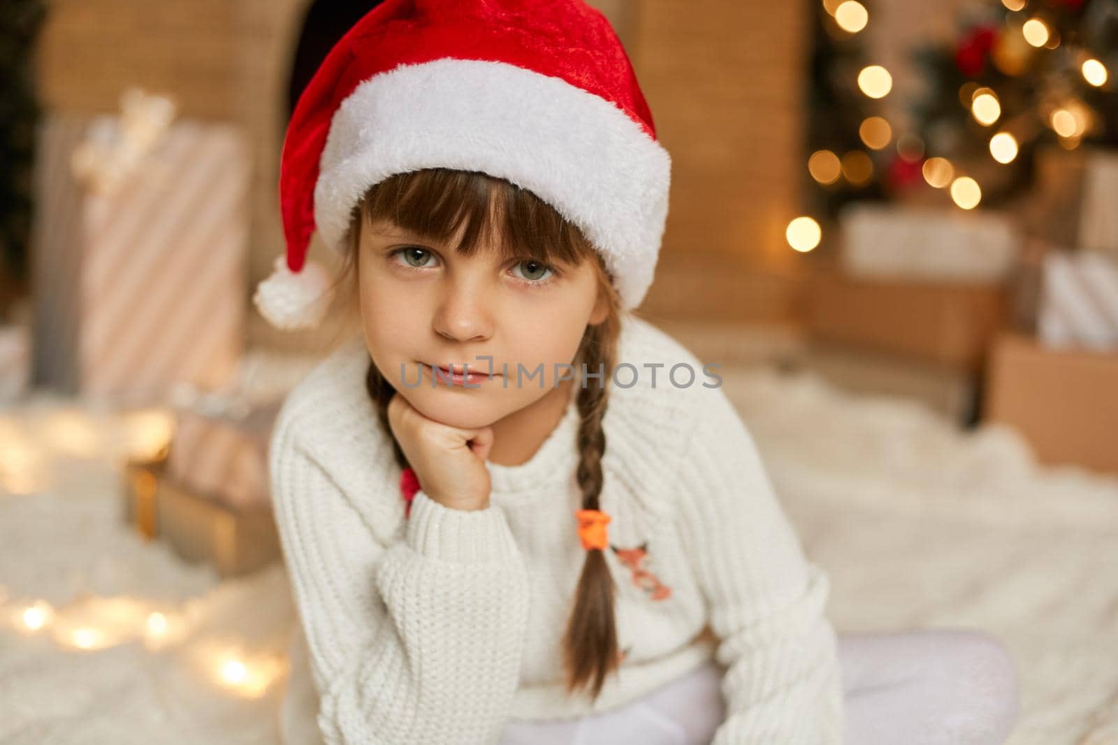 Adorable pensive female child waiting for Christmas, little girl in santa hat wearing white jumper, looks at camera, keeps hand under chin, posing in festive room.