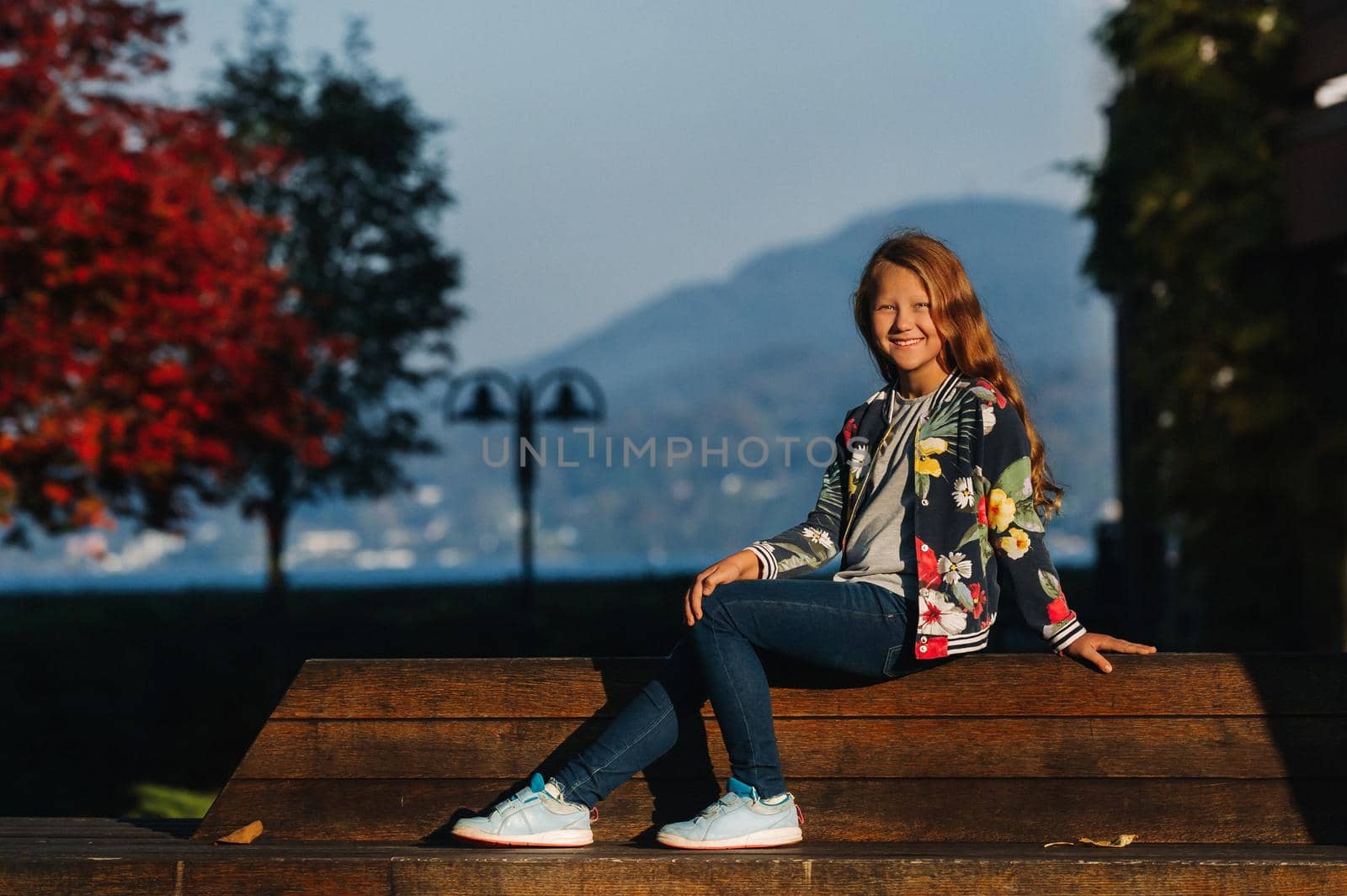 A happy little girl sits on a bench in a Sunny autumn sunset in the old town of Austria. A girl poses in a town against the backdrop of the Austrian Alps.Europe.Velden am Worther See