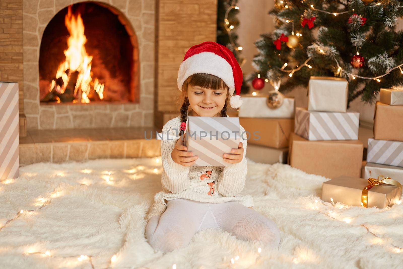 Happy child girl with christmas present at home sitting on warm carpet and looking at box in her hands, has satisfied facial expression, posing near fireplace and Christmas tree.