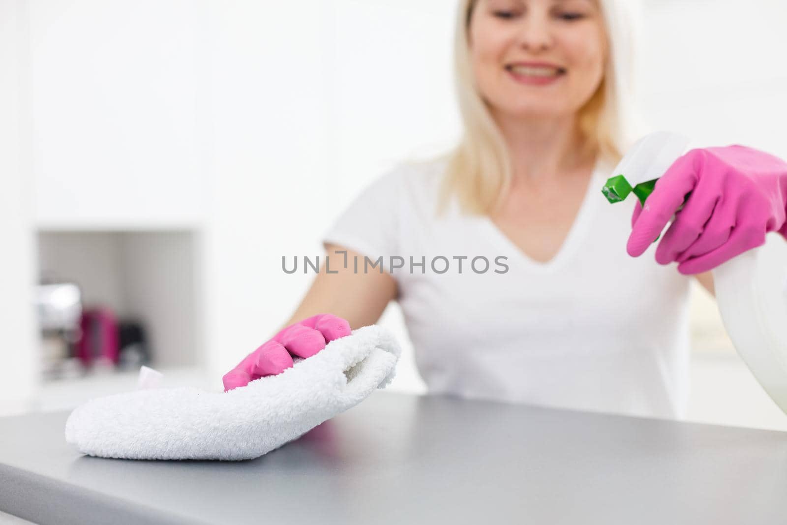 Woman in the kitchen is smiling and wiping dust using a spray and a duster while cleaning her house, close-up by Andelov13