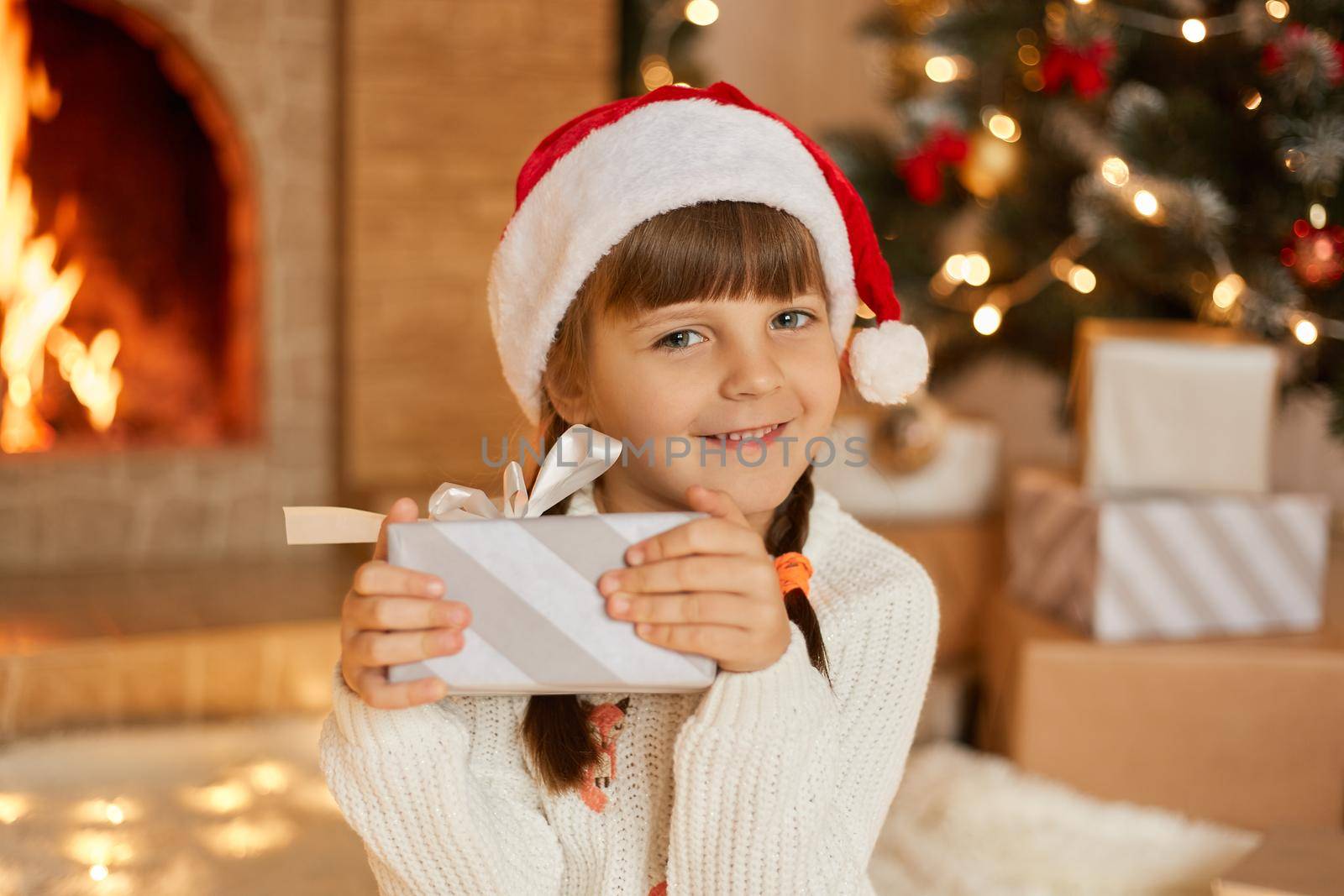 Happy little girl with present at Christmas Eve, sitting near xmas tree and fireplace, charming smiling child looking at camera and holding striped gift box with bow. by sementsovalesia