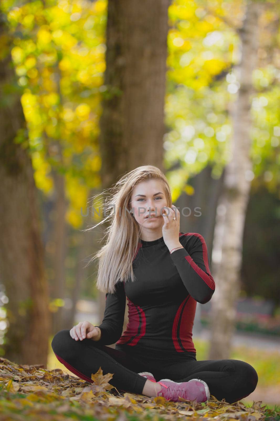 Young attractive fitness female sitting cross-legged at workout in autumn park, straightens her waving in the wind hair, telephoto
