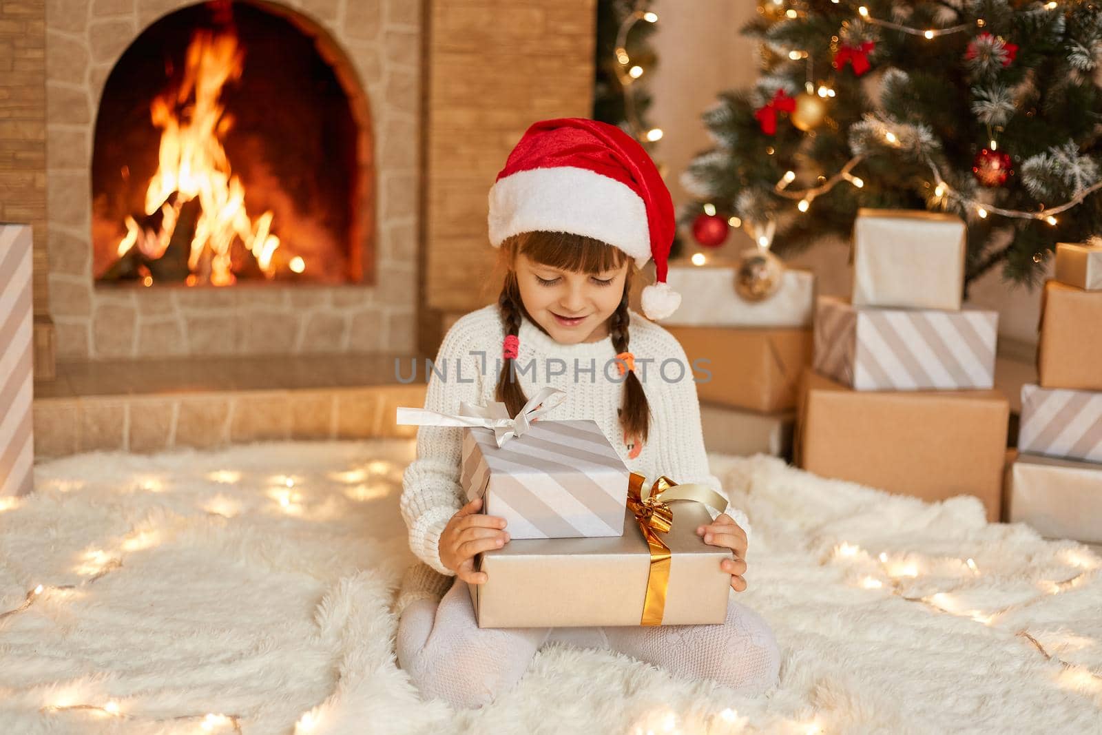 Happy little child by fireplace on Christmas eve sitting on floor on soft carpet with two present boxes, looking at her gifts with happy expression, wants opening them, wearing casual clothing and hat