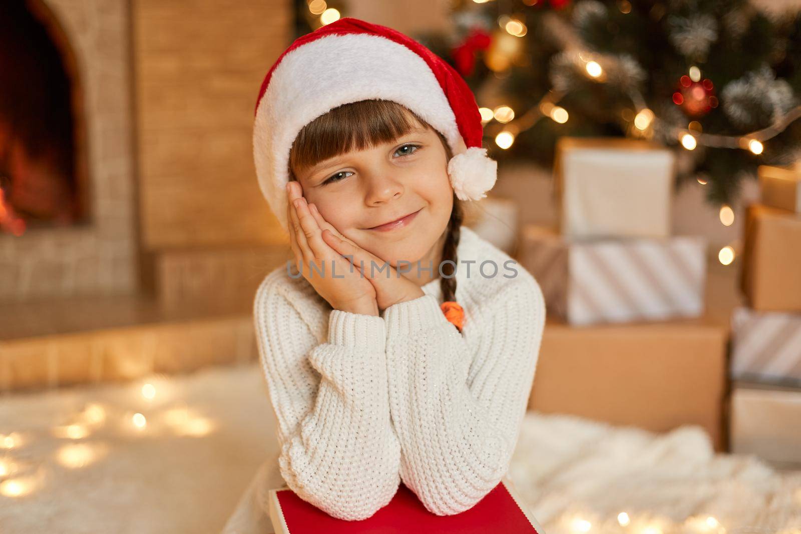 Cute little girl with Christmas present sitting in festive living room, keeping palms together near cheek, looking at camera with satisfied facial expression, being glad to get xmas gifts.