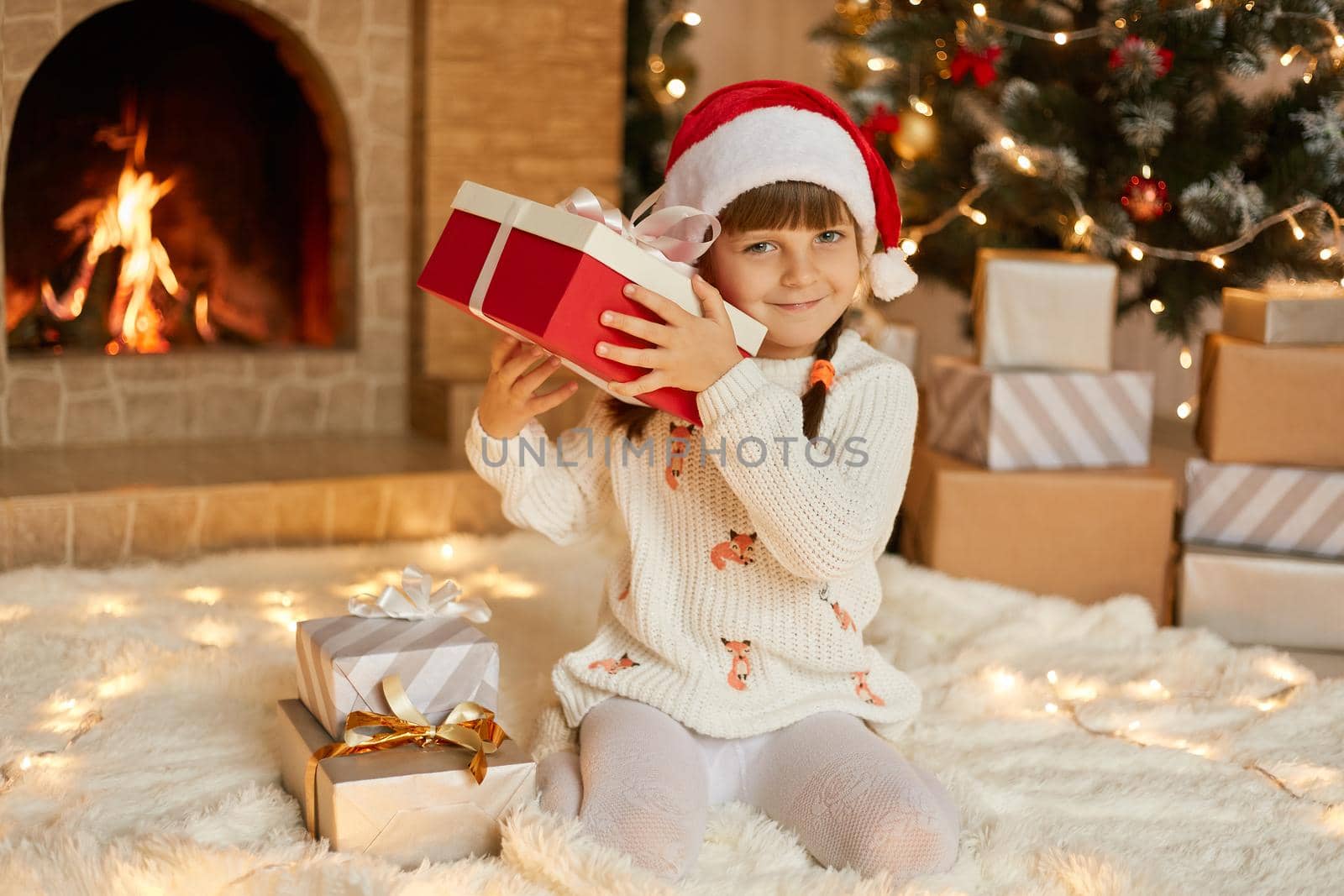 Female child in red hat and warm white jumper sitting on floor in living room with Christmas decoration, shaking gift box , trying to guess what is inside, looks smiling at camera.
