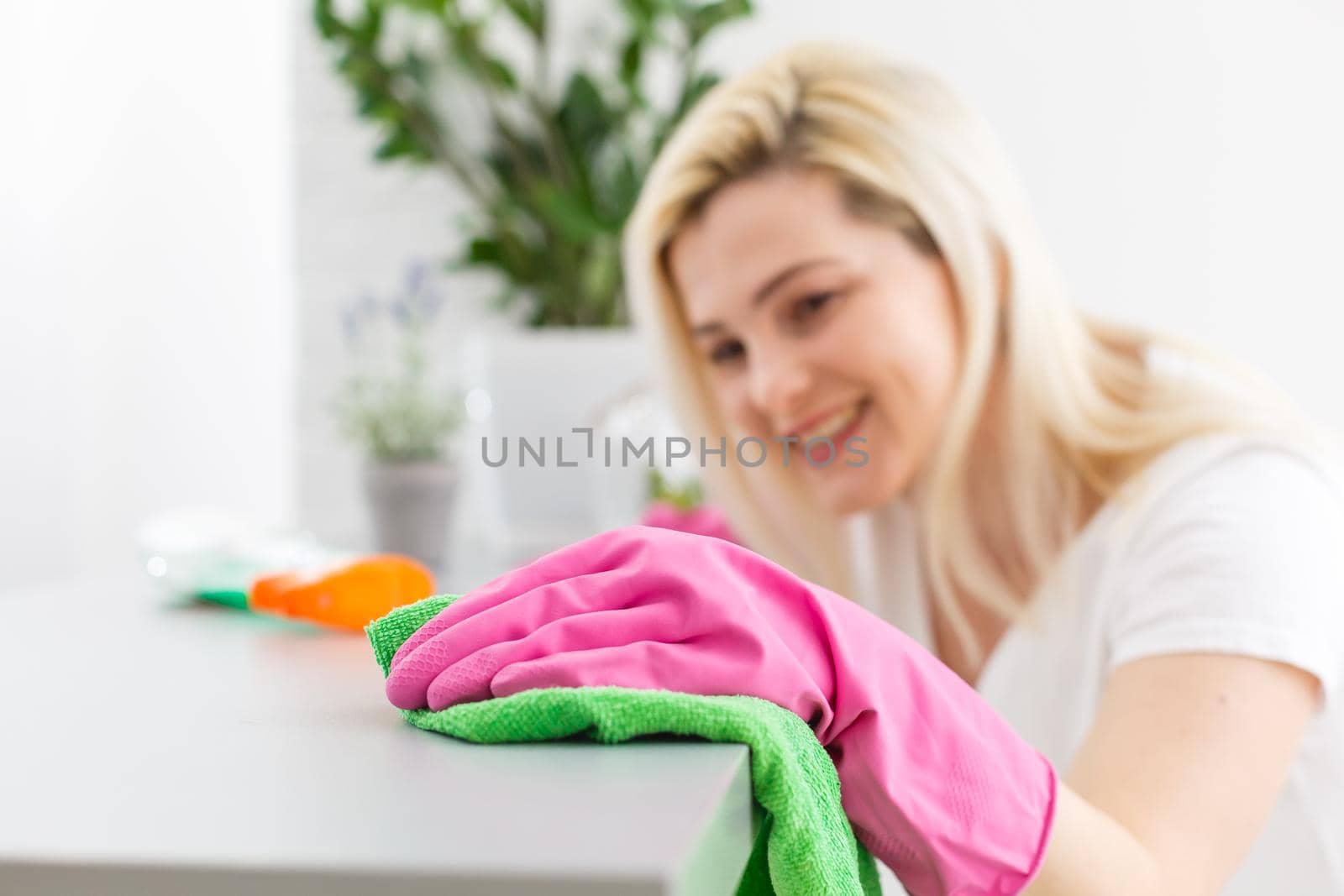 Woman in protective gloves is smiling and wiping dust using a spray and a duster while cleaning her house, close-up by Andelov13