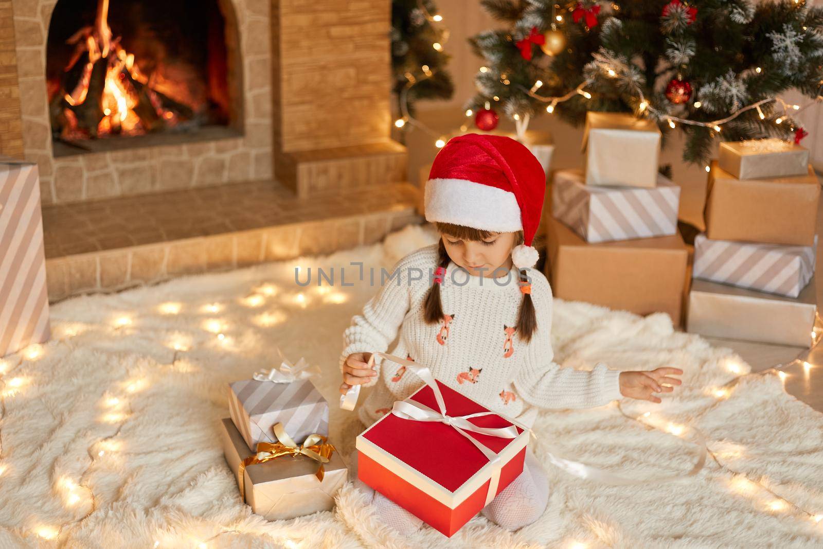 Cute little girl opening Christmas gift at home while sitting on soft carpet on floor, looking at box, pulls ribbon to open present, dresses white pullover and red santa hat.