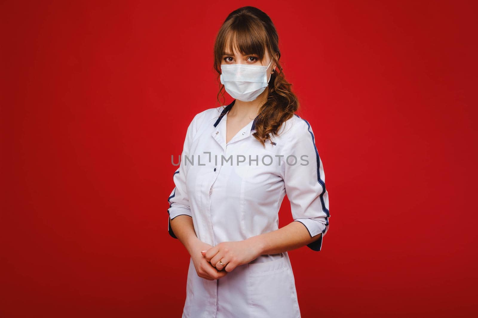 A female doctor stands in a medical mask isolated on a red background. by Lobachad