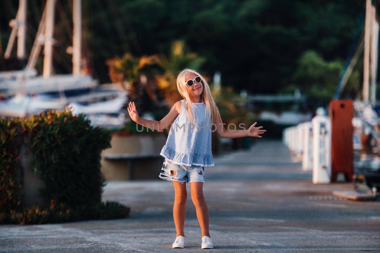 Cute girl near the yachts in the summer. Travel, adventure, boat trips with a child for a family vacation. Children's clothing in the style of a sailor, marine fashion. by Lobachad