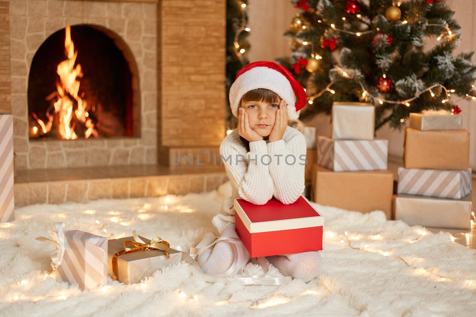 Charming girl sitting on floor with present box, keeping palms on cheeks, looks bored, waiting for opening gifts, posing with xmas tree and fireplace on background, wears santa hat.