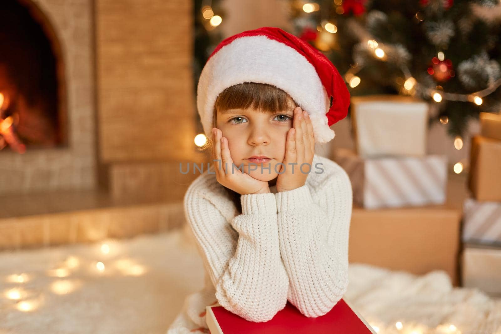 Little girl wearing Santa hat and white sweater sitting near christmas tree and stacks of present boxes with pensive facial expressions, looks at camera, keeps hands under chin.