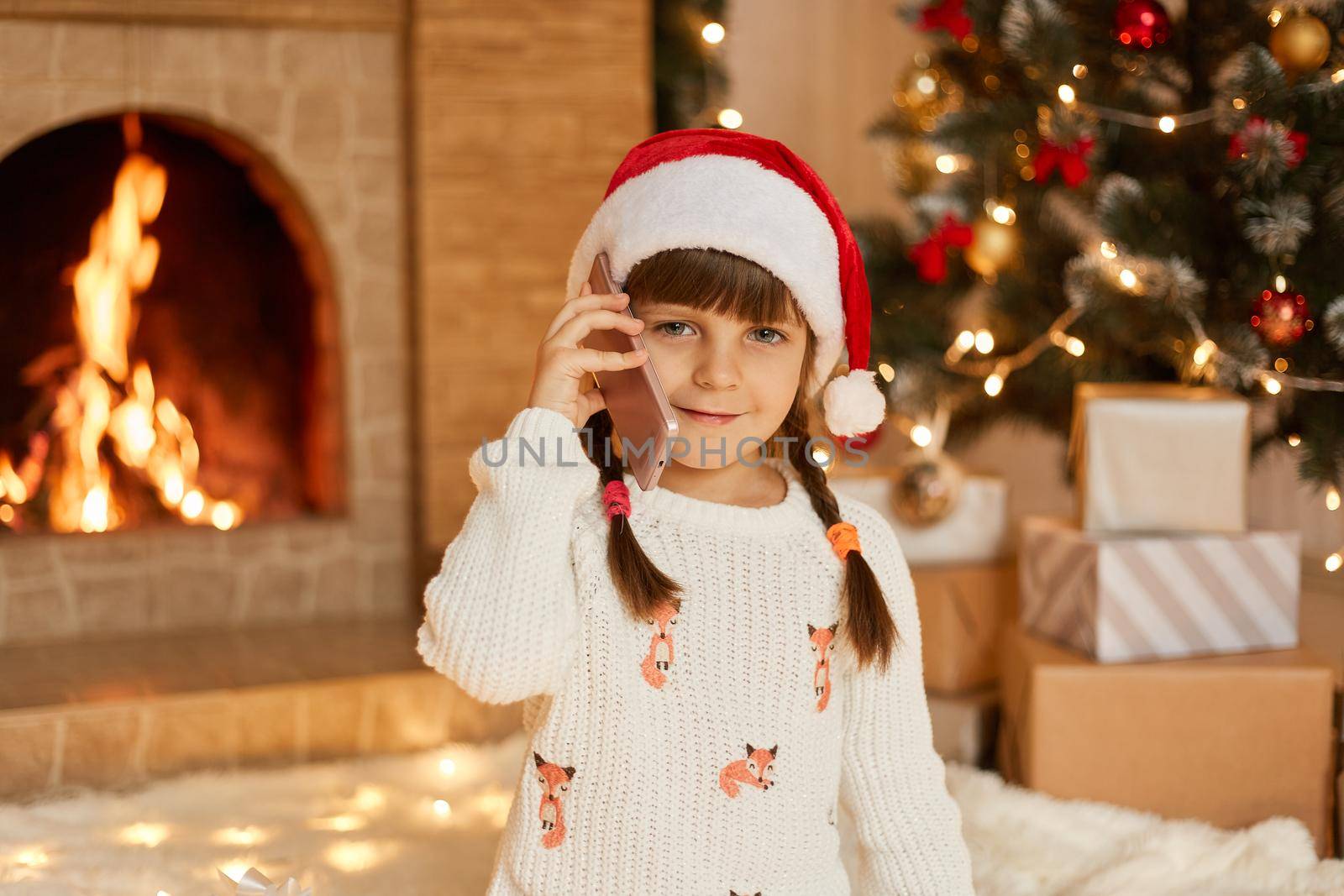 Charming good looking female child talking on phone to somebody, congratulating, having two pigtails, wearing santa hat, looks at camera while posing in living room with Christmas decoration.
