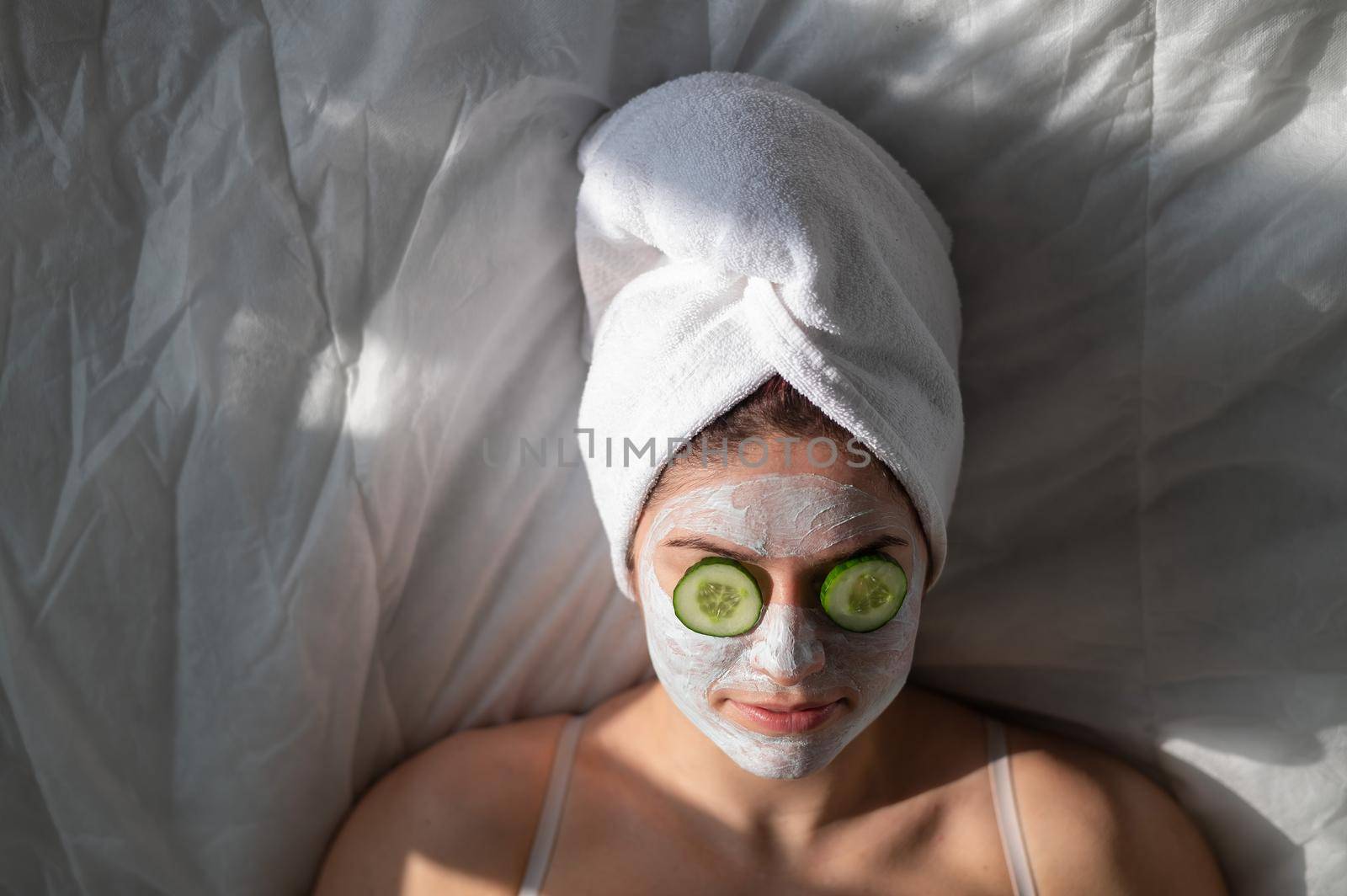 A woman with a towel on her hair and in a clay face mask and cucumbers in front of her eyes lies on the sheet.